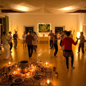 Eine Gruppe von Menschen in einem warm erleuchteten Raum tanzt barfuß in einem Kreis um einen kunstvollen Kakaozeremonie-Altar mit Kerzen, Tonkrügen und einer Lotusblumen-Dekoration im Hintergrund